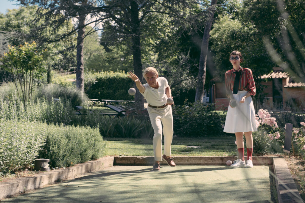 Due persone giocano a bocce su un piccolo campo immerso nel verde di un giardino alberato. Un uomo lancia la boccia con gesto concentrato, mentre una donna accanto a lui osserva la traiettoria tenendo in mano un’altra boccia. Intorno, siepi curate, fiori e alberi creano un’atmosfera rilassata e conviviale all’aria aperta.