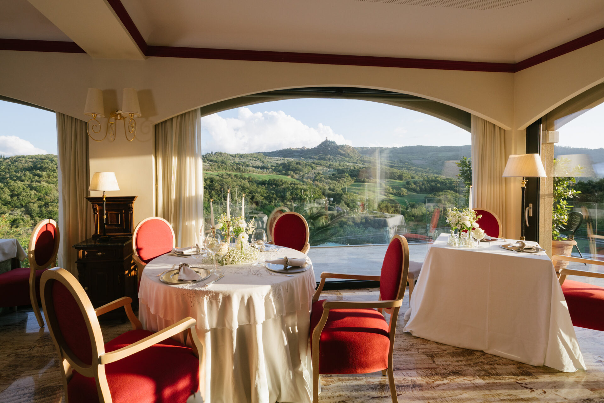 Elegante sala ristorante con ampie vetrate ad arco che incorniciano un panorama mozzafiato sulle colline verdi. Tavoli rotondi e quadrati vestiti di tovaglie bianche sono apparecchiati con cura, impreziositi da composizioni floreali delicate. Le sedie imbottite rosse aggiungono un tocco classico e raffinato all’ambiente, illuminato dalla luce calda del sole che entra dalle finestre. Atmosfera romantica e panoramica, ideale per una cena esclusiva.