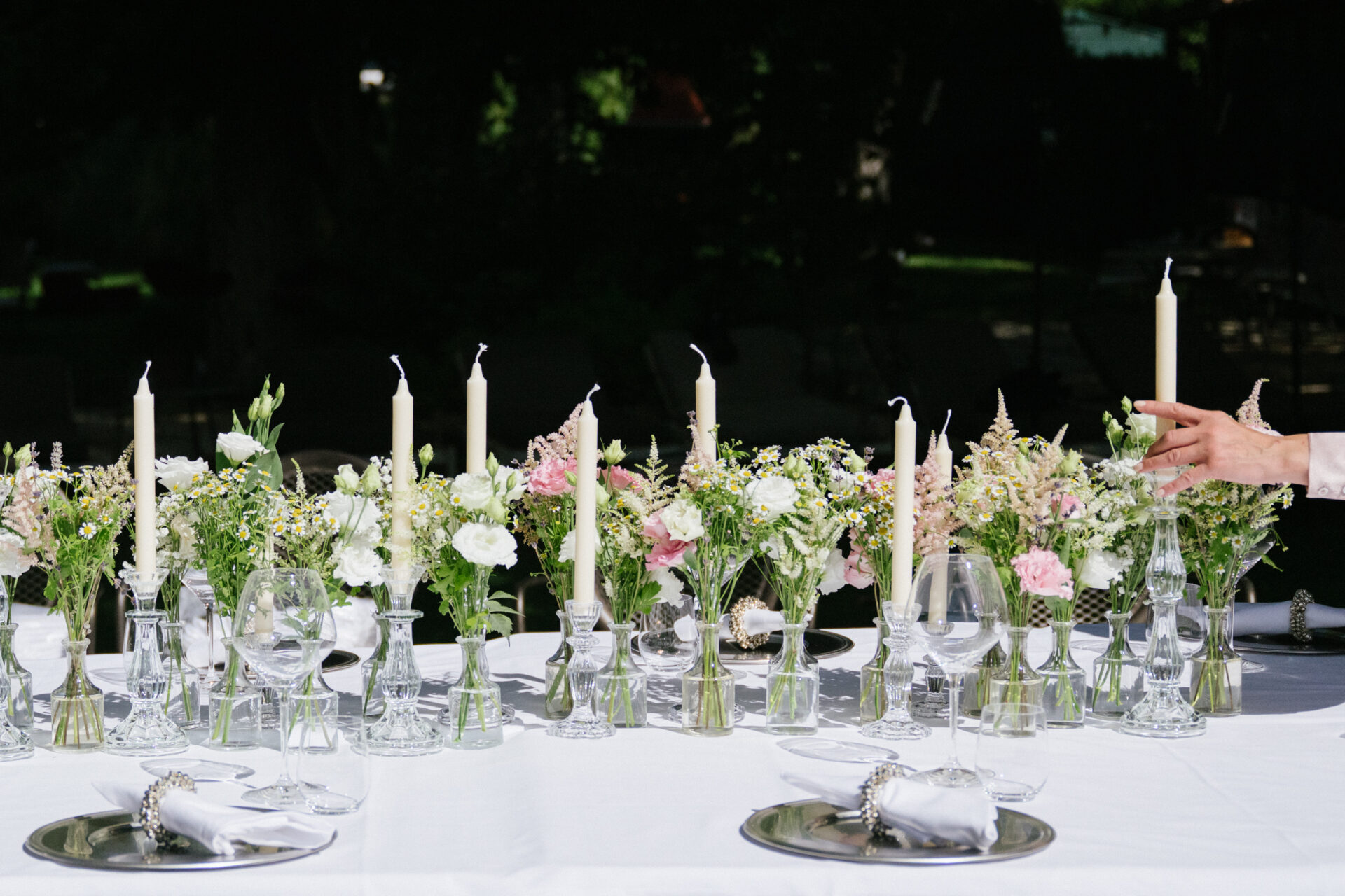 Tavola elegantemente apparecchiata con calici in cristallo, piatti con sottopiatti argentati e candele bianche disposte tra composizioni floreali fresche nei toni del bianco, rosa e verde. Una mano sistema i dettagli dell’allestimento, creando un’atmosfera raffinata e romantica, ideale per un evento o una cena speciale.