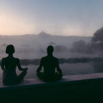 Due persone sedute in posizione meditativa sul bordo di una piscina termale avvolta dalla foschia del mattino. Le loro silhouette si stagliano contro il paesaggio collinare della Val d’Orcia all’alba, con ombrelloni e profili di alberi sullo sfondo. Atmosfera silenziosa, contemplativa e profondamente rilassante.