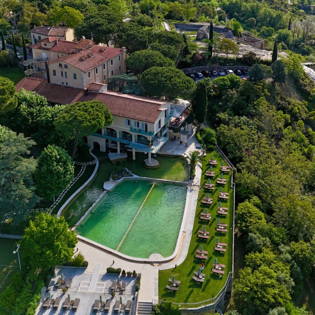 Vista aerea di un hotel immerso nel verde con piscina termale rettangolare color smeraldo, solarium con lettini ordinati sul prato e terrazze affacciate sulla vallata. Intorno, alberi rigogliosi e colline toscane creano un’atmosfera di relax e armonia con la natura.